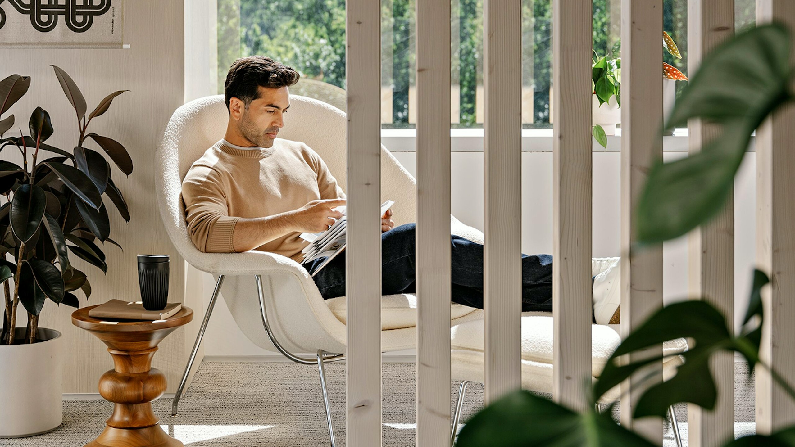 A person with short brown hair, wearing a beige sweater and blue jeans, sitting in a white Knoll Womb Chair and Ottoman while reading next to green potted plants