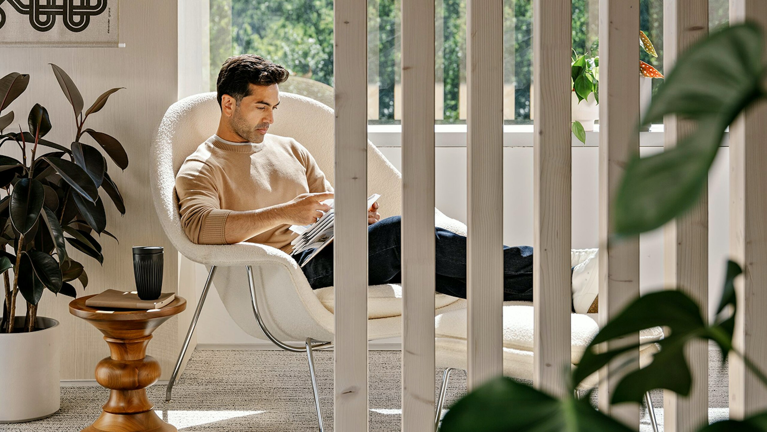 A person with short brown hair, wearing a beige sweater and blue jeans, sitting in a white Knoll Womb Chair and Ottoman while reading next to green potted plants