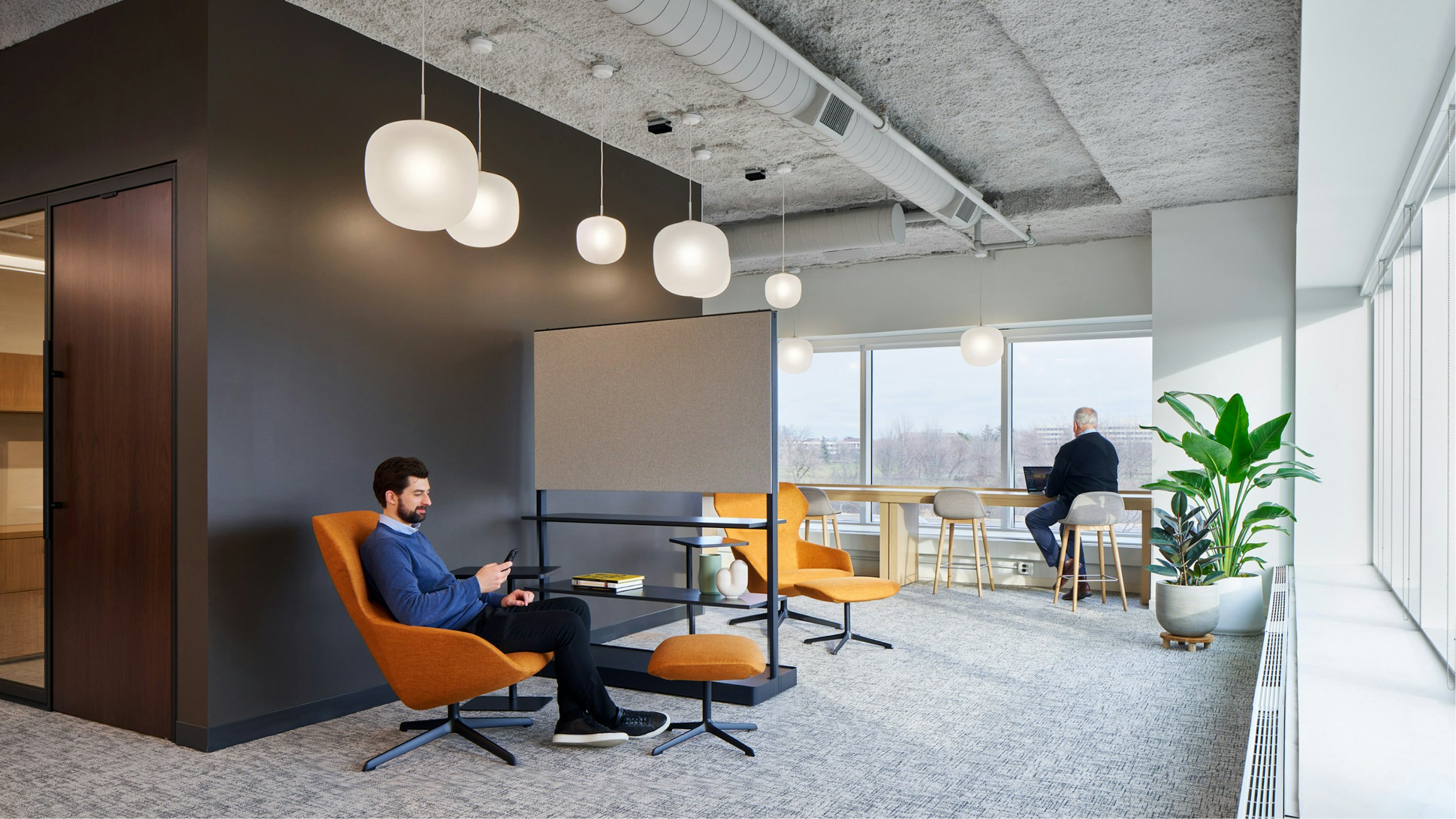 A person in a blue sweater sitting in an orange lounge chair and ottoman while another person works at a bar height counter in the office of an industrial organization