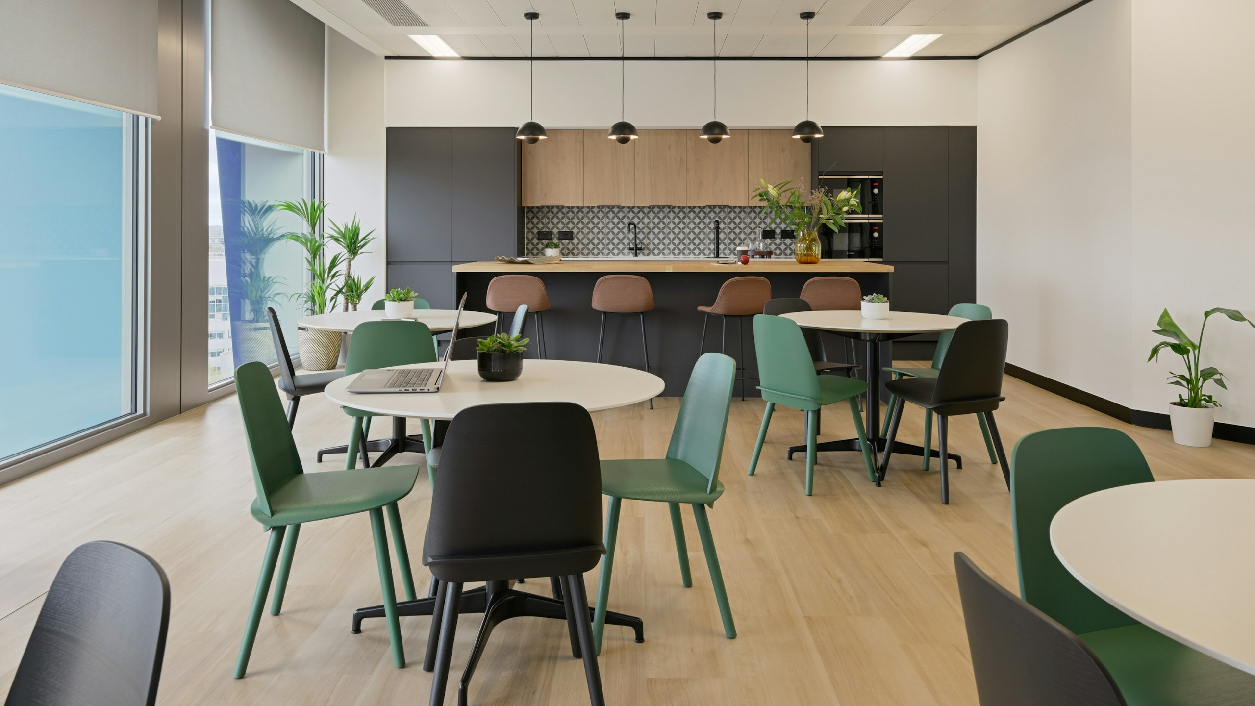 A communal kitchen space at the NATO Defence Innovation Accelerator in London furnished with green and black chairs, round Civic tables, and bar-height stools at a kitchen island