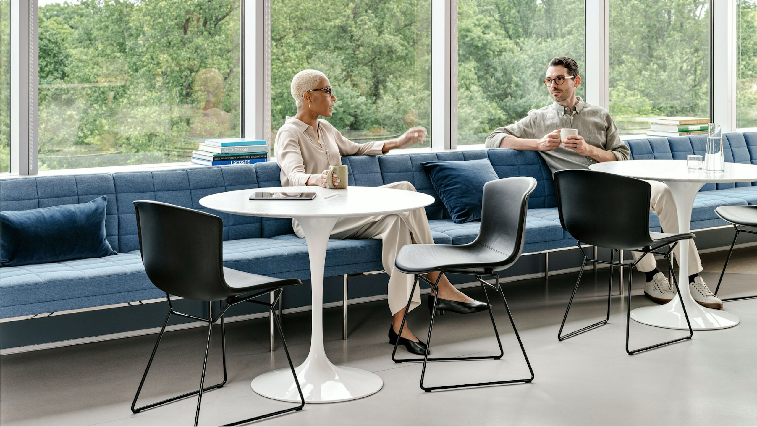 Two individuals conversing in a shared seating area furnished with Saarinen Tables, Knoll Bertoia chairs, and blue bench seating